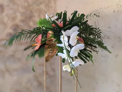 Composition florale exotique sur colonne dorée avec orchidées blanches, anthuriums roses et feuillage tropical