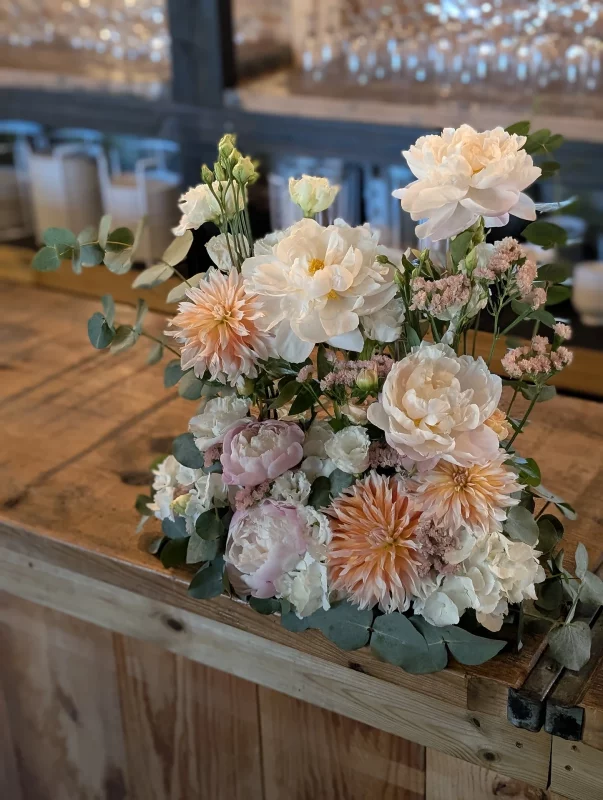 Classic wedding floral arrangement with peonies, peach dahlias, roses and eucalyptus on a wooden bar.