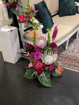 Grand arrangement floral avec proteas roses, anthuriums rouges, celosia et feuillages tropicaux, ambiance exotique.