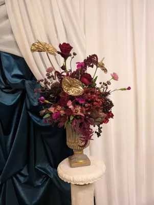 Impressionnante composition florale classique avec roses bordeaux, anthuriums dorés et orchidées magenta, posée sur un piédestal élégant.