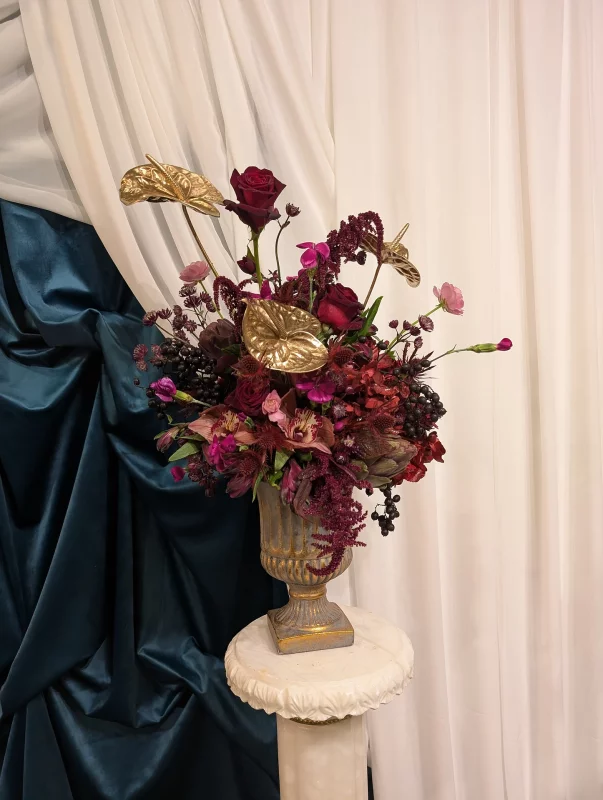 Impressionnante composition florale classique avec roses bordeaux, anthuriums dorés et orchidées magenta, posée sur un piédestal élégant.