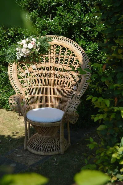 Scénographie de cérémonie boheme avec composition florale de roses et eucalyptus sur chaise paon en rotin pour mariage.