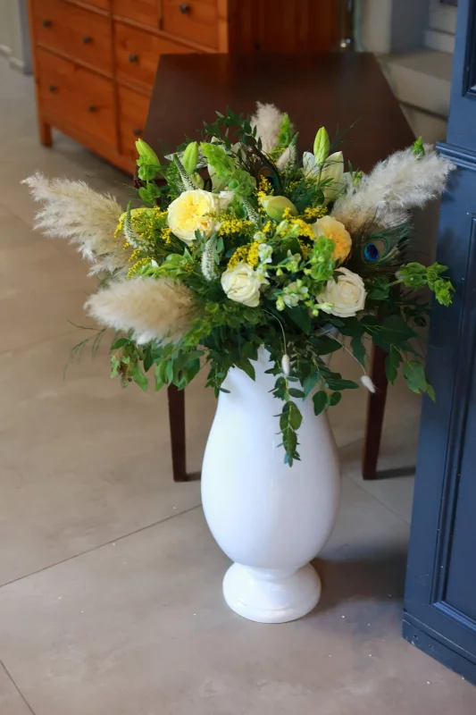 Grand bohemian floral composition in a white vase, featuring yellow roses, pampas grass and a peacock feather.