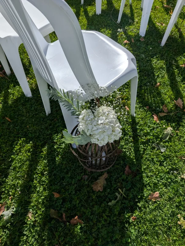 Countryside Ceremony Aisle Decoration featuring white hydrangeas, gypsophila, and lush foliage, presented within a rustic metal cage, positioned at ground level.