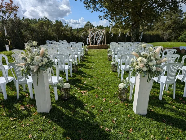 Décoration d'allée de cérémonie bohème avec des compositions florales de pampas, roses blanches et eucalyptus sur des socles en bois pour un mariage en extérieur.