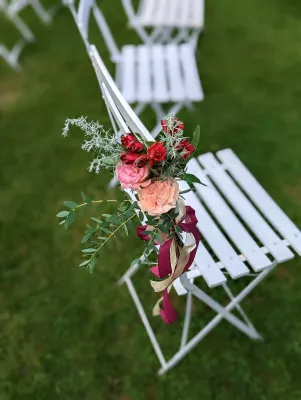 Décoration de chaise de cérémonie avec un petit bouquet de roses rouges, roses et pêche, feuillage et rubans bordeaux et beige, style champêtre.