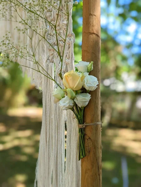 Detail de decoration de ceremonie boheme avec roses blanches, gypsophile et feuillage vert sur poteau en bois.