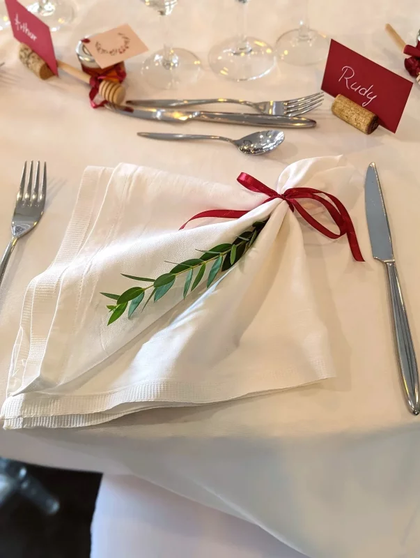 Décoration simple de serviette de mariage avec ruban bordeaux et brin de feuillage vert, élégance minimaliste pour les invités.