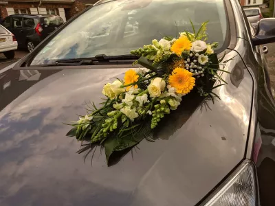 Décoration de véhicule de mariage champêtre avec gerberas jaunes, roses blanches et feuillage verdoyant, pour un jour spécial.