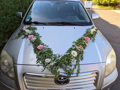 Décoration florale de voiture de mariés avec guirlande de feuillage, roses pâles et gypsophile pour un style champêtre.