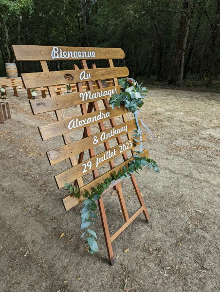 Panneau de bienvenue en bois champêtre pour mariage, orné d'une guirlande d'eucalyptus et d'une composition florale rouge et blanche.