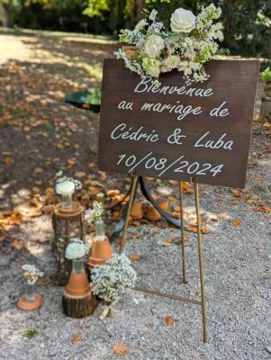Panneau de bienvenue en bois pour mariage, décoré de roses blanches et gypsophile, avec buches et pots en terre cuite.
