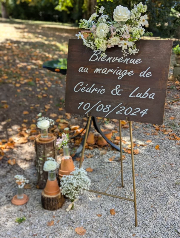 Wooden wedding welcome sign, adorned with white roses and gypsophila, featuring logs and terracotta pots.