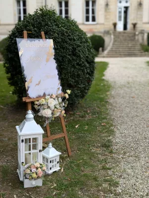 Scénographie d'accueil de mariage avec panneau de bienvenue, composition florale de roses, œillets et lanternes blanches.