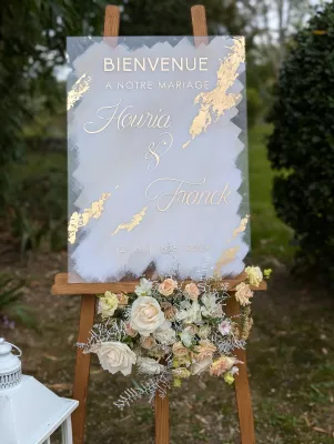 Gros plan sur un panneau de bienvenue de mariage personnalisé, avec arrangements floraux de roses et feuillage argenté.