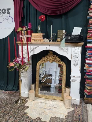 Scénographie de mariage avec cheminée décorative, livres anciens, miroir doré 'Bienvenue', et chandelier floral, style classique bohème.