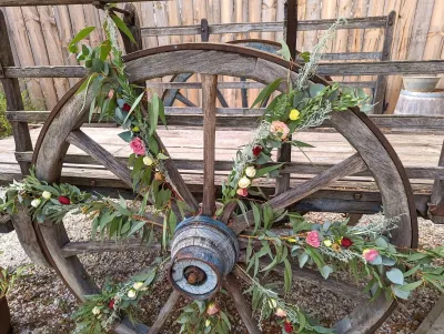 Scénographie de mariage champêtre avec une ancienne roue de charrette ornée d'une guirlande d'eucalyptus et de petites roses pour un coin photo.