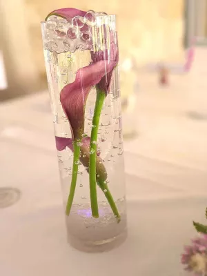 Détail d'un centre de table de mariage avec un vase cylindrique en verre, des callas violets immergés et des billes de gel.