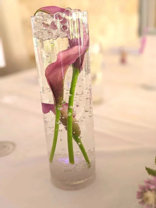 Détail d'un centre de table de mariage avec un vase cylindrique en verre, des callas violets immergés et des billes de gel.