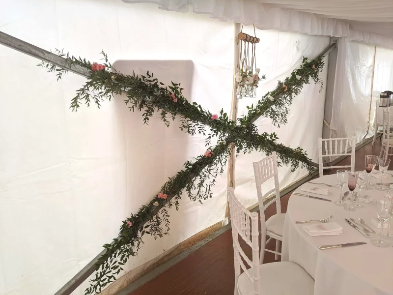 Végétalisation murale en forme de croix avec du ruscus, du lierre et de petites roses roses pour une décoration de salle.